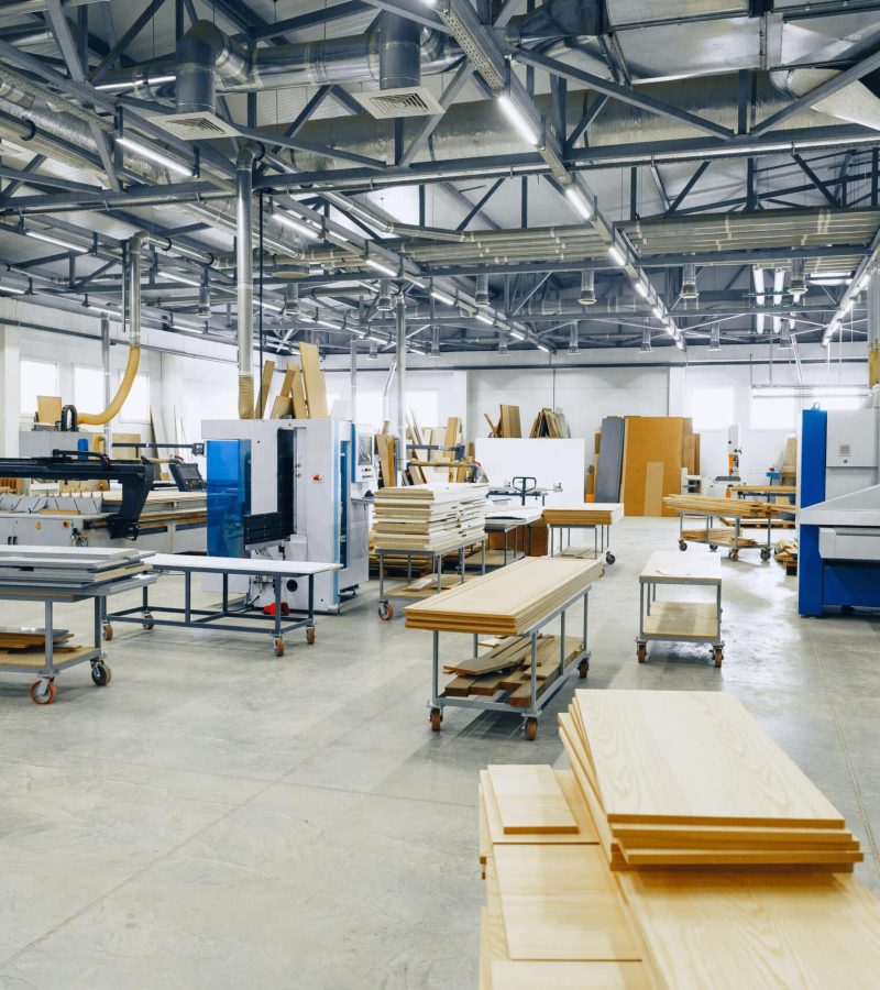 Workers use machines and tools in a large furniture workshop with many pieces of wood.